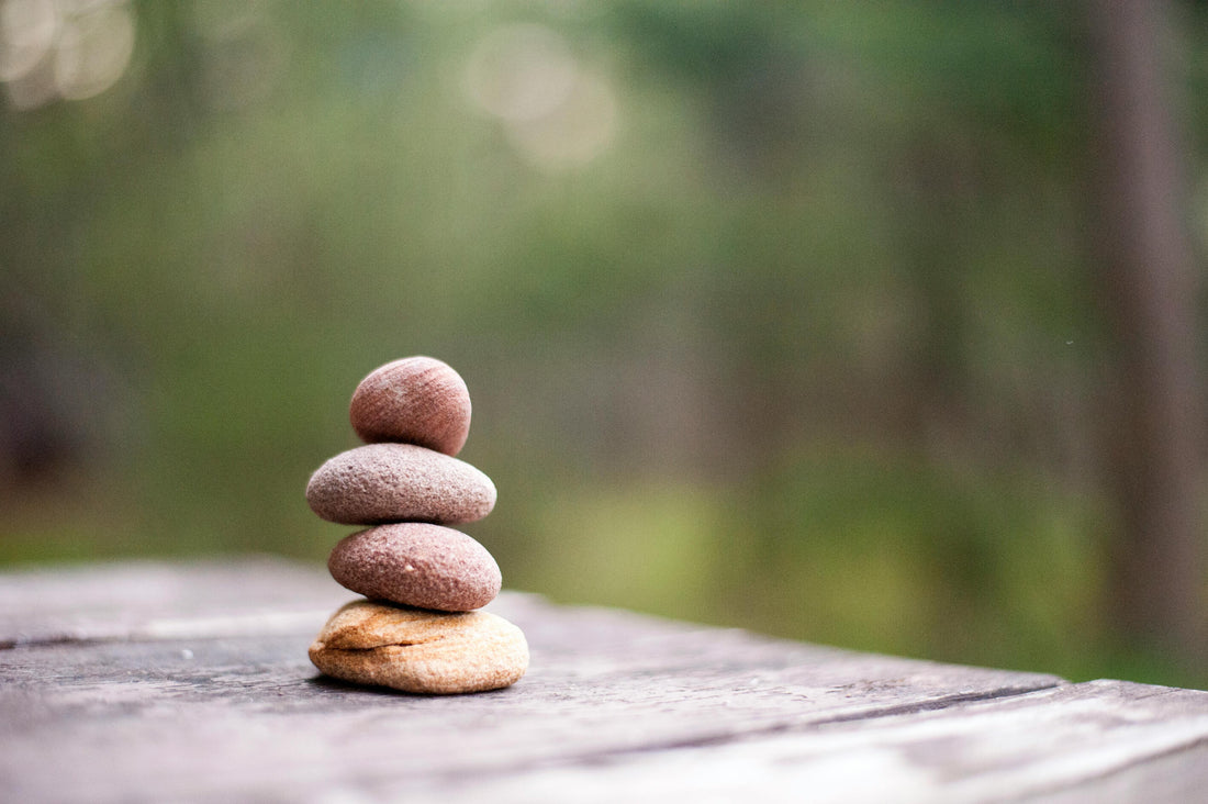A photo of 4 river rocks stacked together on a wooden floor. The camera is focused on the rocks, and in the background are some blurred trees.